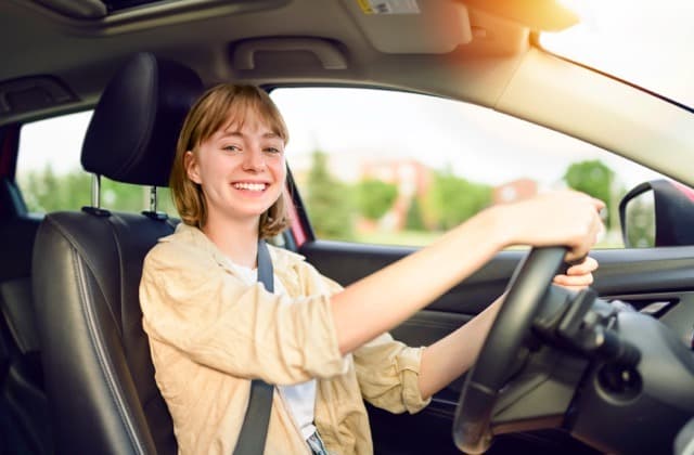 A smiling young woman sitting in the driver's seat of a car with both hands on the steering wheel.