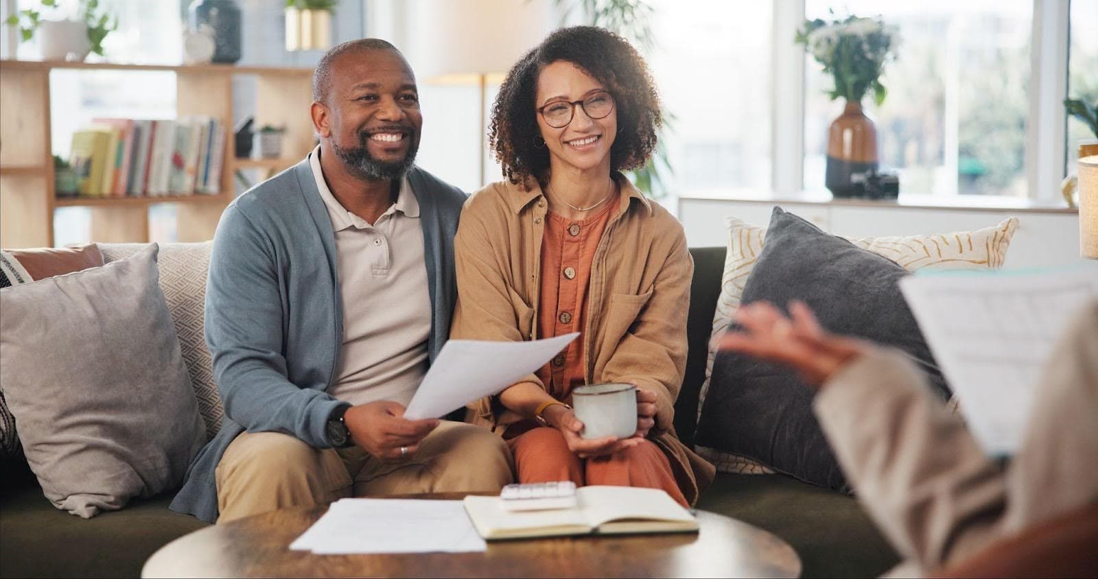 A couple reviews a life insurance policy together on a couch, holding a cup of coffee and discussing their options.