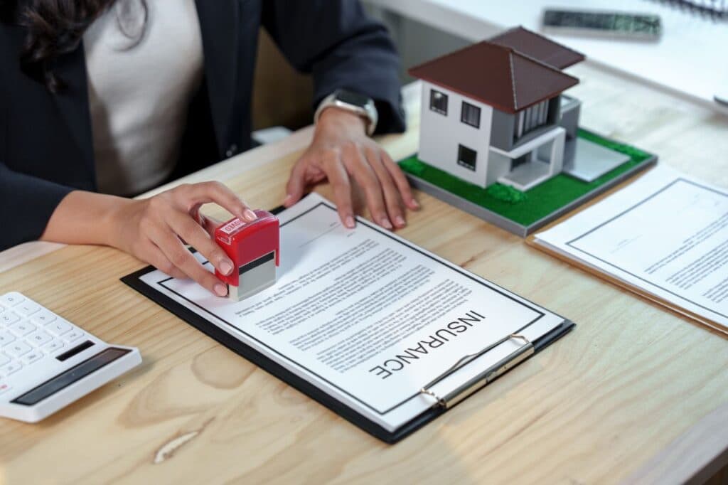 Person stamping "APPROVED" on home insurance document with house model and calculator on desk.