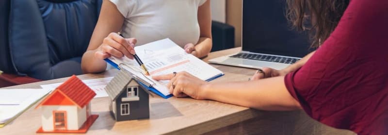 A woman is signing documents while a small model house is placed on the table beside her, symbolizing a real estate transaction.