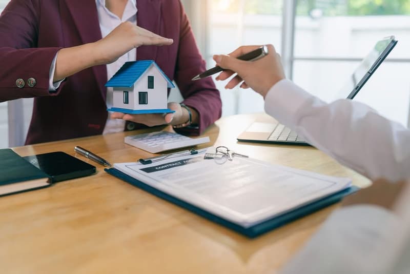 Real estate agent protecting a model house with hands above it during a client consultation.